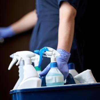 A cleaner wearing dark blue scrubs and gloves carries a bucket with spray bottles