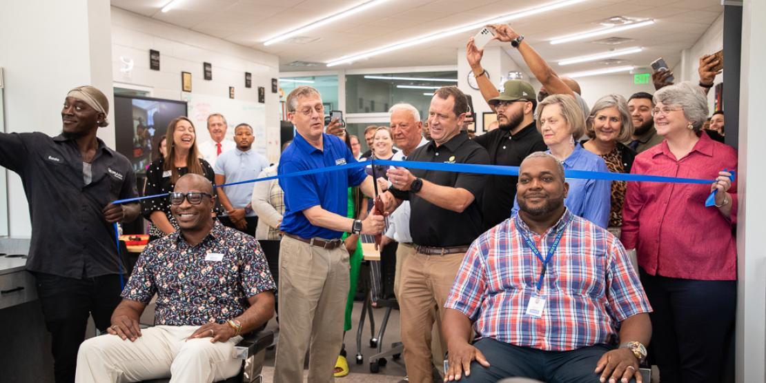 People gather in renovated barbershop as a blue ribbon is cut with oversized scissors 